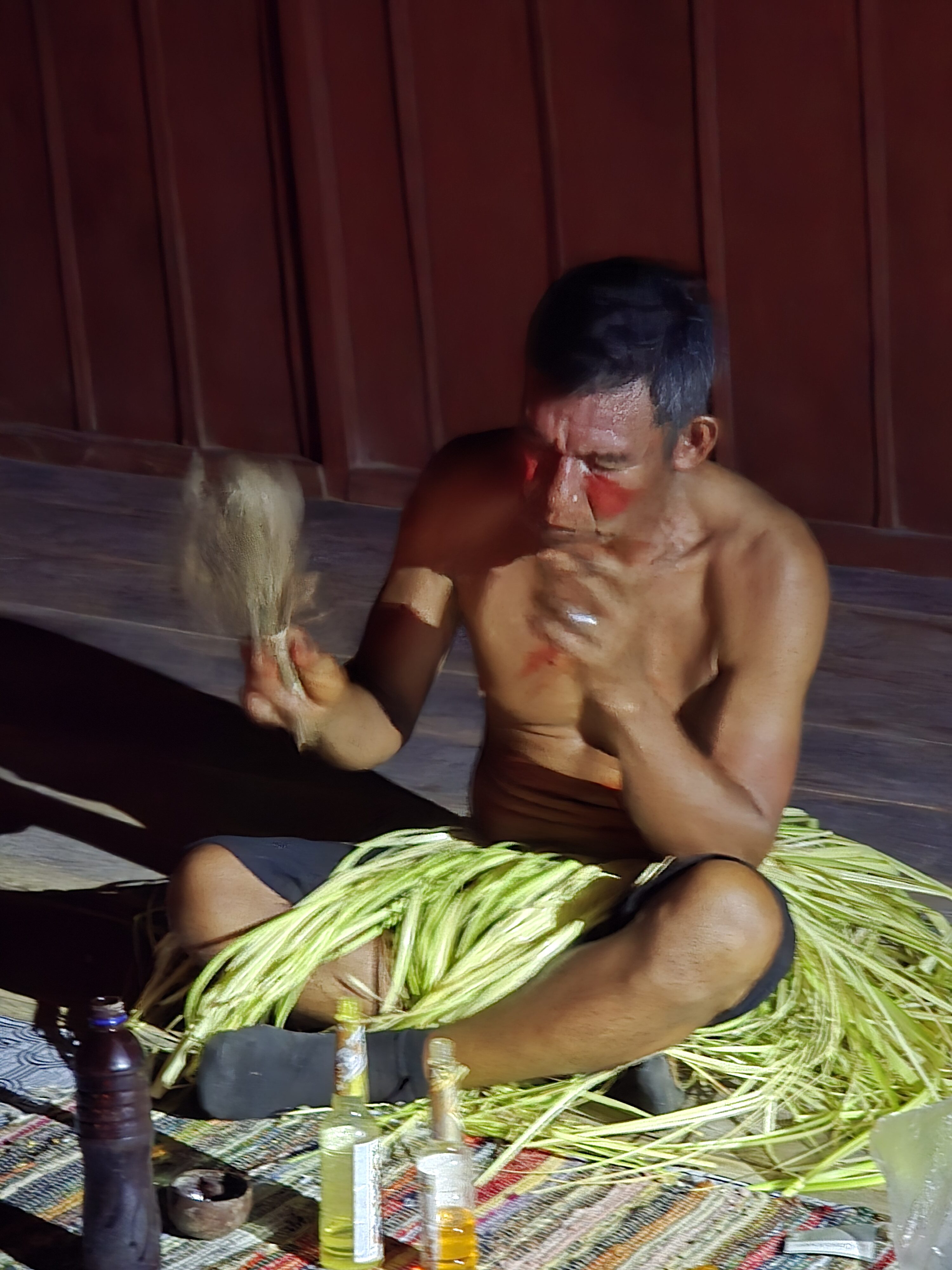 A local guide with traditional red face paint sits on a woven mat inside a wooden pavilion at Selva Wild Lodge, demonstrating a cultural ritual using palm fibers and natural materials while bottles and small containers are arranged in front of him.